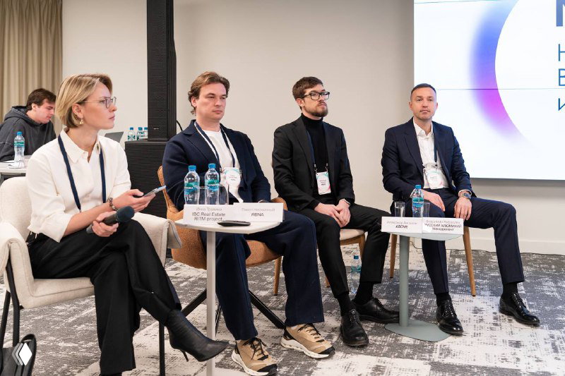 Панельные спикеры сидят за столом на сцене форума Next Point: модератор и четыре участника обсуждают новые подходы к проектированию офисов