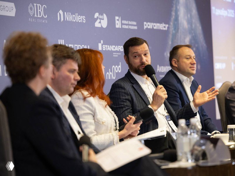 Панельная дискуссия: несколько экспертов коммерческой недвижимости беседуют за столом, один из спикеров держит микрофон; сцена с брендированием мероприятия.