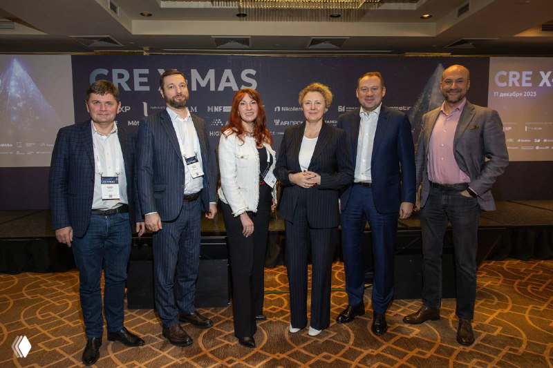 Групповая фотография спикеров и организаторов на фоне баннера CRE X‑MAS 2025: участники конференции стоят в деловой одежде на сцене в конференц‑зале.
