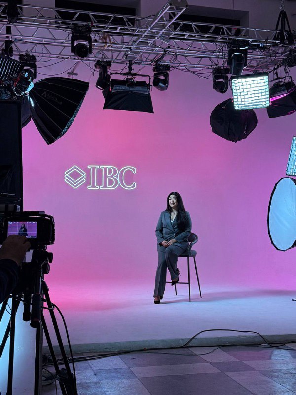 Женщина сидит на стуле в студии на розовом фоне; за ней логотип IBC, вокруг видны камеры и световое оборудование.