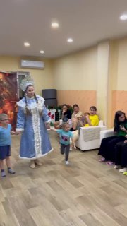 Снегурочка в движении среди детей в гостиной приюта: малыши бегают и улыбаются, взрослые наблюдают и поддерживают праздник.
