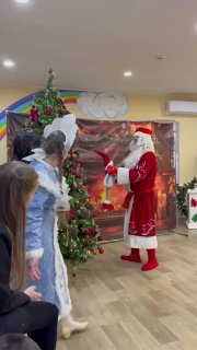 Дед Мороз возле елки показывает что‑то детям на фоне праздничного декора — сцена праздничного представления в приюте.