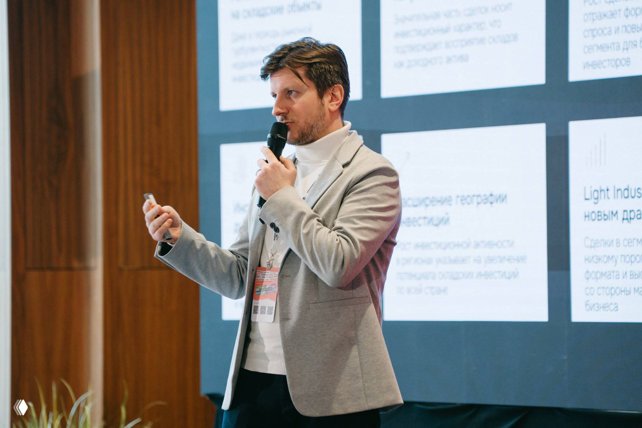 Спикер в светлом пиджаке проводит презентацию у большого экрана с инфографикой; в кадре видно пульт и значки участника на бейдже, актовый зал заднего плана.
