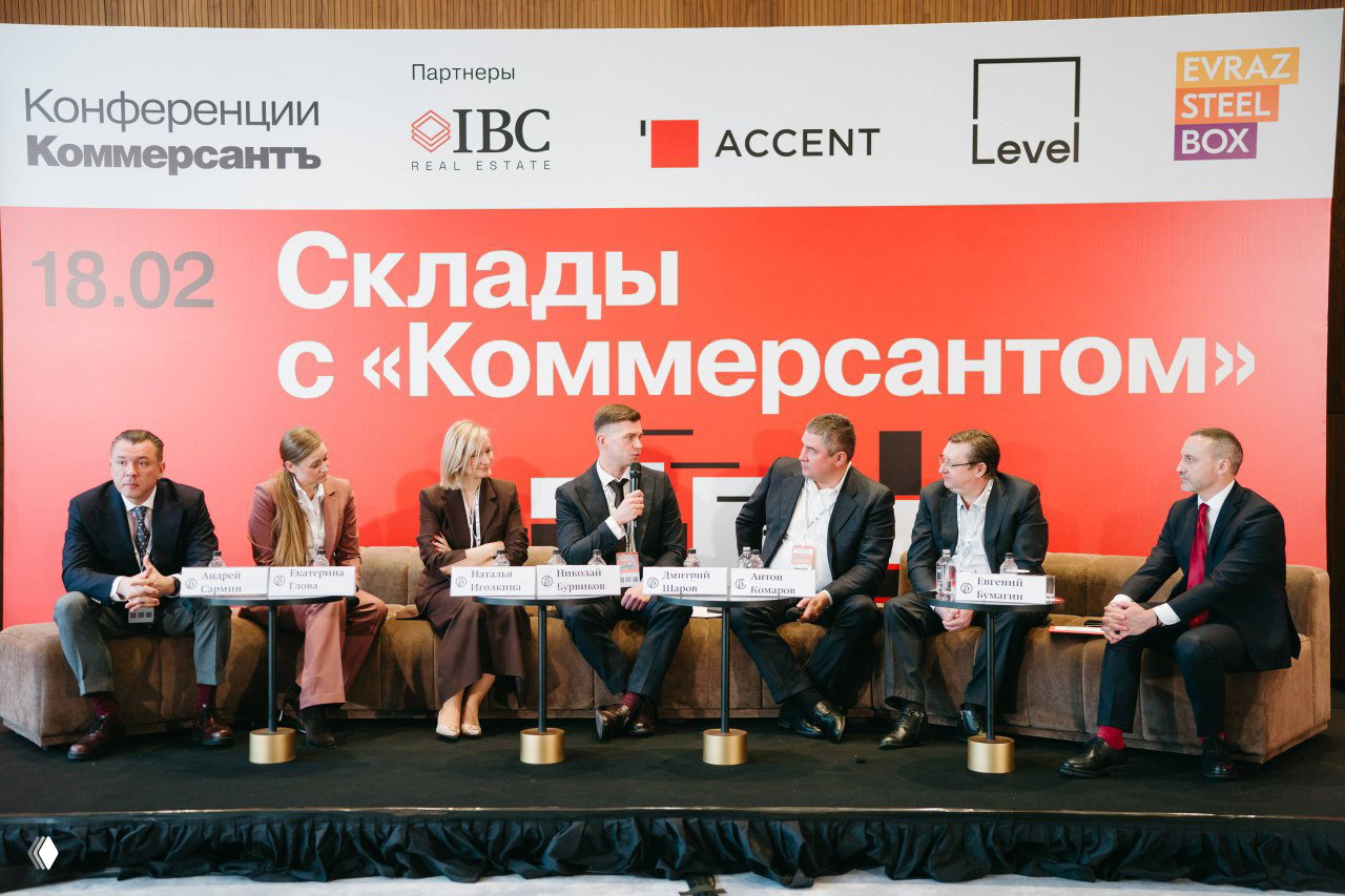 Панельная дискуссия на сцене: несколько спикеров сидят на диванах на фоне яркого баннера с надписью конференции «Склады с "Коммерсантом"», аудитория вне кадра.