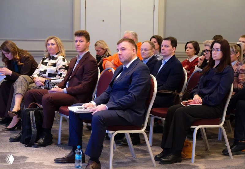 Зрители и участники конференции IBC Real Estate слушают доклад, сидя в зале: деловые костюмы, раздаточные материалы и внимание к спикеру.