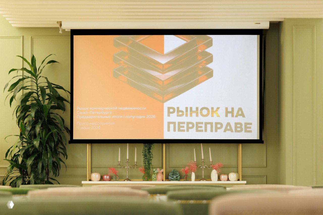 Проекционный экран с оформлением и заголовком «Рынок на переправе» в зале пресс‑завтрака IBC Real Estate, вид на декор и сцену мероприятия.
