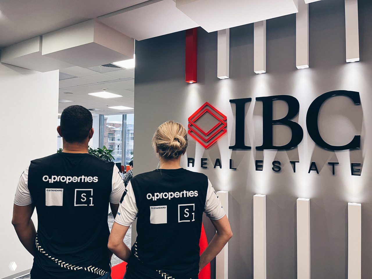 Двое человек в майках o.properties стоят спиной в офисе рядом с крупным логотипом IBC Real Estate — командный корпоративный кадр.
