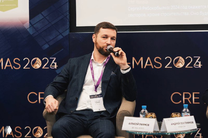 Представитель IBC Real Estate выступает в микрофон на панельной сессии CRE X-mas, видны стол и таблички спикеров.