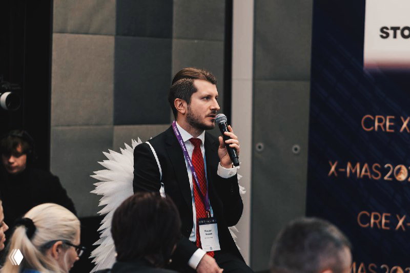 Спикер IBC Real Estate говорит в микрофон перед аудиторией на CRE X-mas; на фоне видно брендирование мероприятия и слушателей.