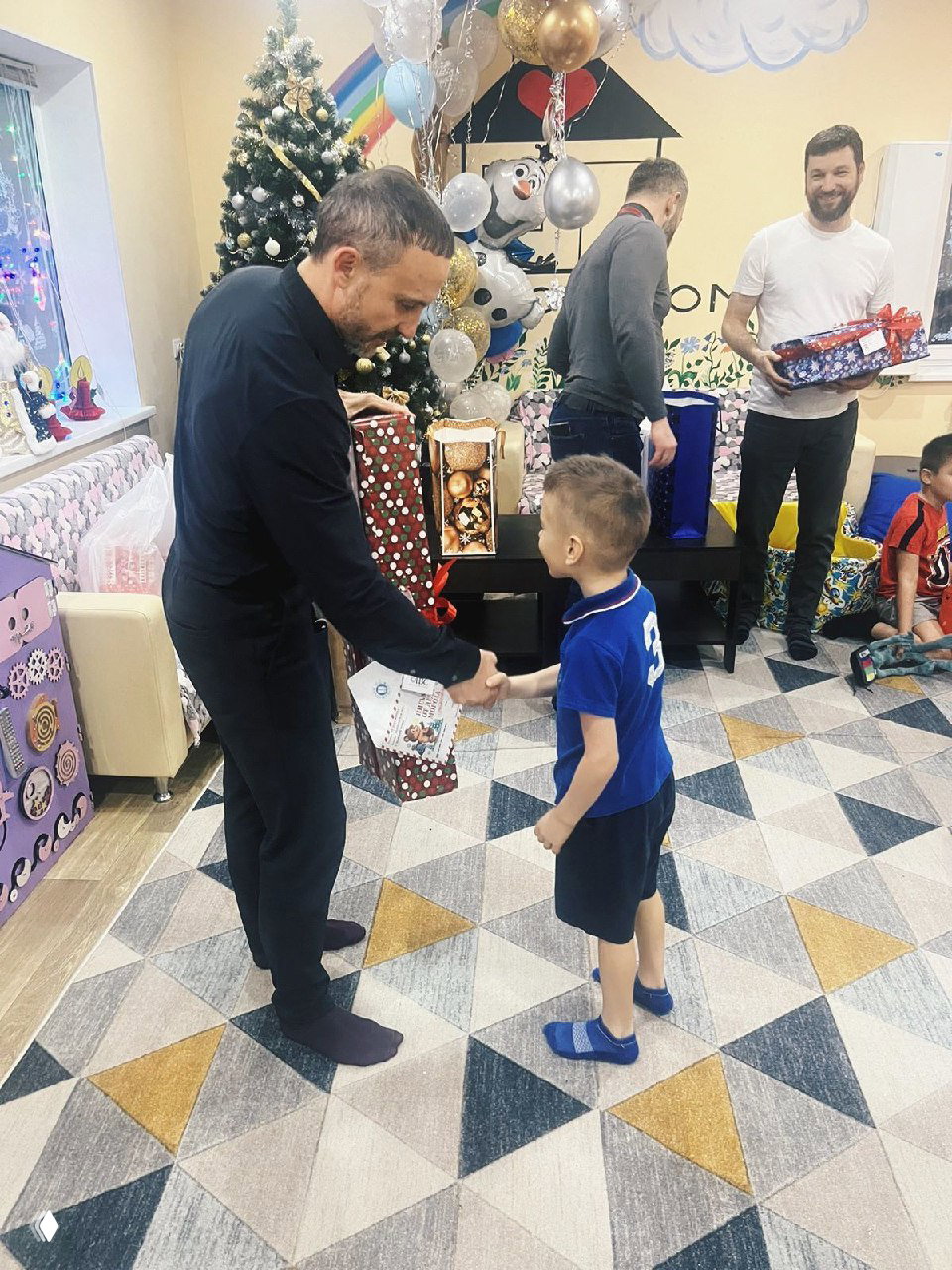 Момент передачи подарка: сотрудник пожимает руку мальчику и вручает коробку, на заднем плане другие участники и праздничные декорации приюта.
