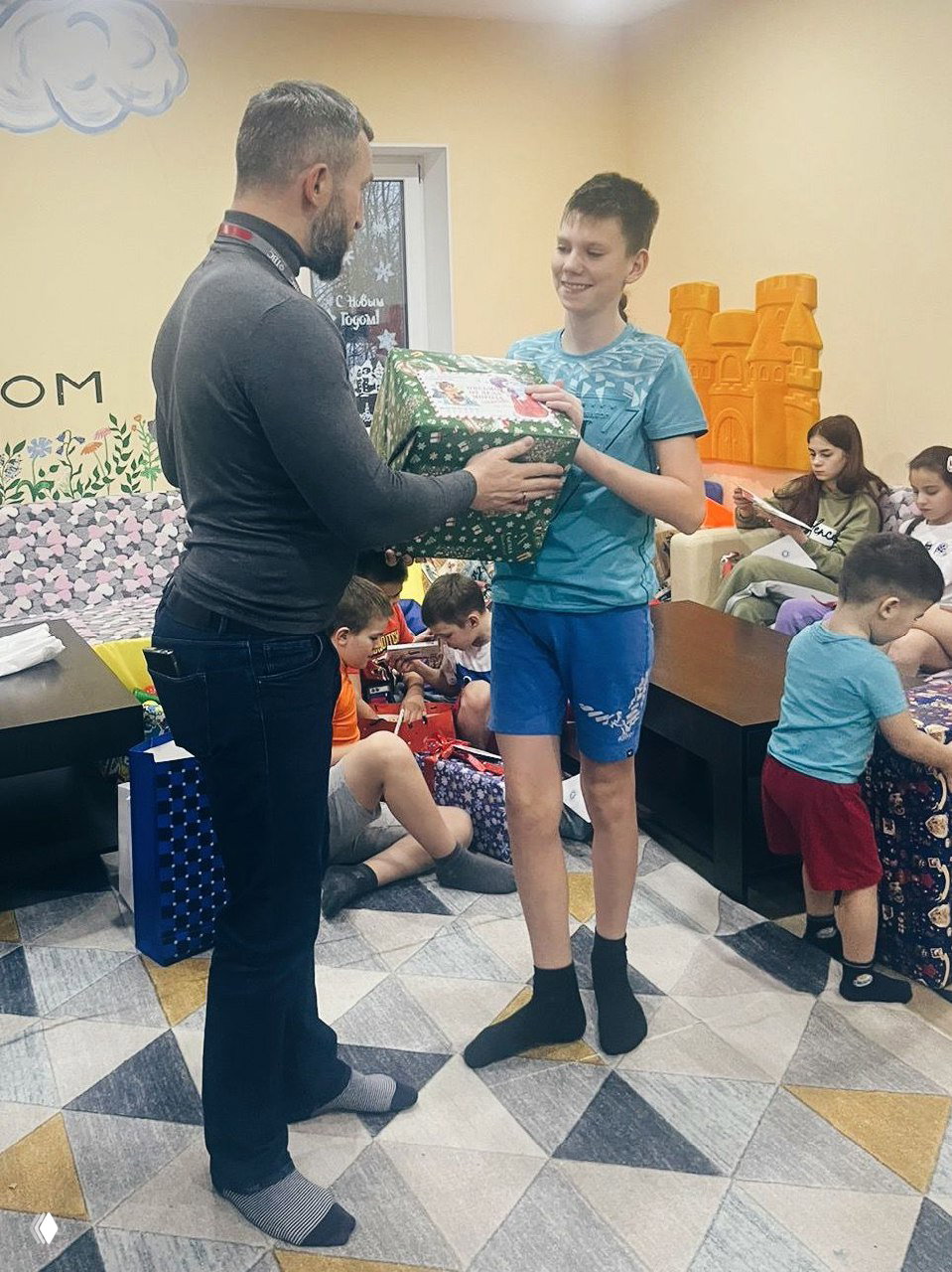 Вручение большого праздничного подарка подростку: сотрудник передаёт коробку высокому воспитаннику, вокруг дети распаковывают свои презенты.