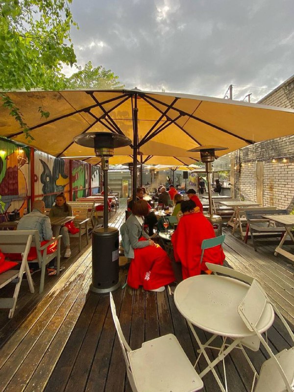 Летняя терраса паба с большими зонтиками, посетители в пледах и уличными обогревателями, деревянные столы и непринуждённая атмосфера на свежем воздухе.