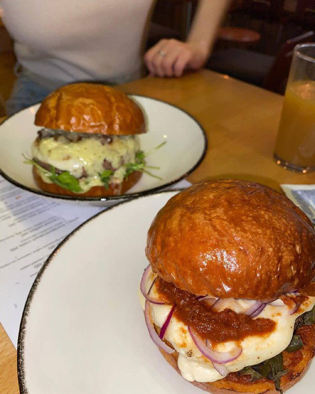 Два бургерa на белых тарелках в интерьере гастробара: глянцевые булочки, сыры и соусы, атмосфера уютного ужина в городе.