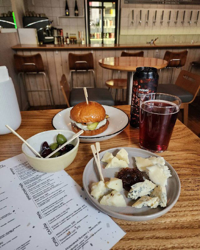 Стол в кафе с мини-бургером, бокалом тёмного напитка и тарелкой закусок; на столе видимо меню — кадр уютной городской трапезы.