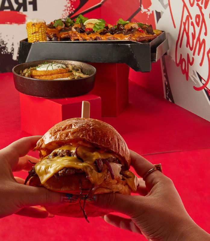 Руки держат большой сочный бургер с тающим сыром и хрустящими элементами на ярком фоне фуд-зоны — крупный и аппетитный кадр.