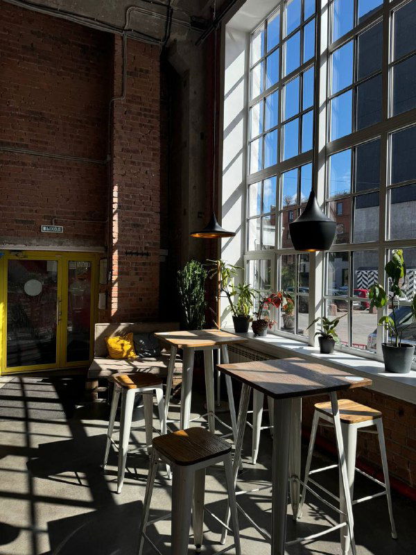 Просторный индустриальный зал кофейни с высокими окнами, деревянными столами и барными стульями — ощущение коворкинга и цеха.