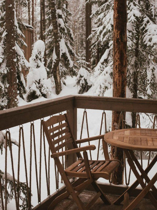 Деревянный балкон с креслом и круглым столиком у заснеженных деревьев: спокойная зимняя терраса загородного домика с видом на лес.