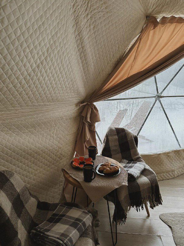 Интерьер купольного глэмпинга: пледы в клетку, небольшой столик со снеками и чашкой на фоне панорамного окна с видом на заснеженный лес.