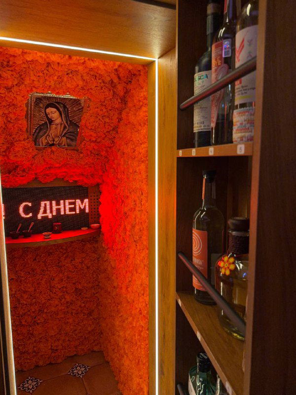 Интерьер мини-мескалерии внутри Paloma Cantina: полки с бутылками, насыщенно-оранжевые стены и неоновая вывеска у входа в закрытую комнату.