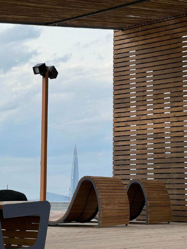 Деревянная беседка и волнообразные лавочки у воды; через проём конструкции вдалеке виднеется шпиль Лахта Центра под облачным небом.