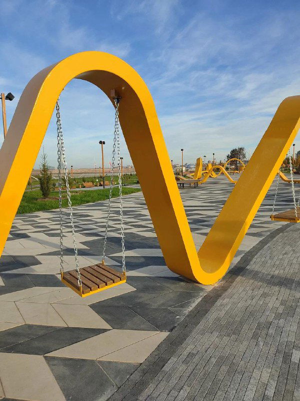 Современная детская зона с яркими желтыми арками и подвесными качелями на благоустроенной набережной в ясный день с голубым небом.