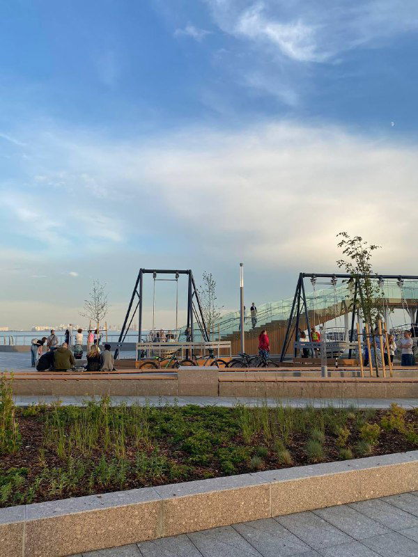 Пространство на набережной с качелями и скамейками, люди прогуливаются у воды; видны низкие бордюры, зелёные насаждения и открытое небо.
