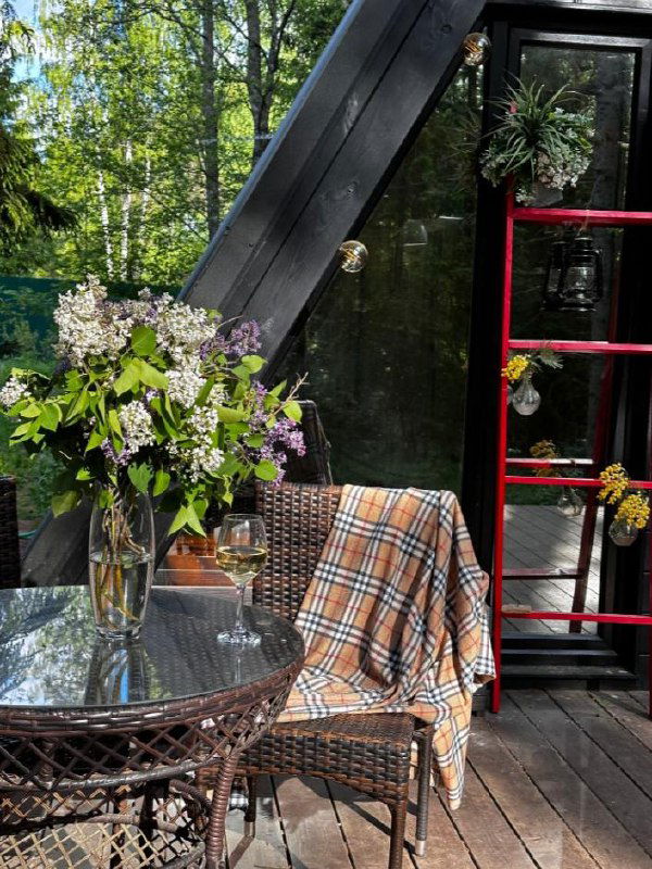 Деревянная терраса с плетёным столом, вазой с цветами и клетчатым пледом; уютный уголок на свежем воздухе среди деревьев.