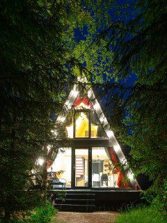 А-образный шале в лесу вечером: тёплый свет из окна, видна внутренняя обстановка и лестница; домик окружён сосновыми стволами.