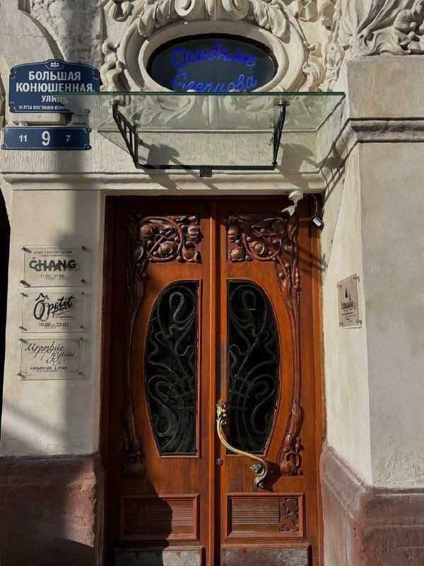 Входные двери Особняка Слепцова на Большой Конюшенной, украшенные резными деревянными створками и табличками кафе и мастерских внутри.