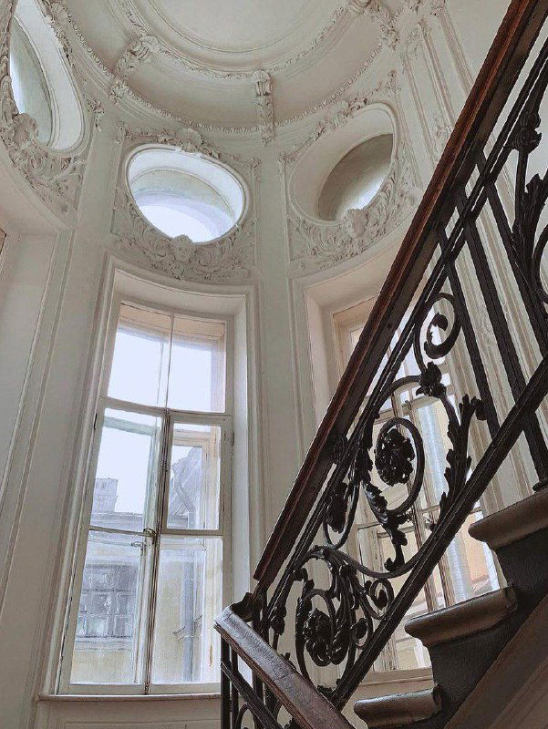 Фото лестницы с коваными перилами и высокими окнами в парадной; типичный ракурс для фотографий в особняке с исторической отделкой.