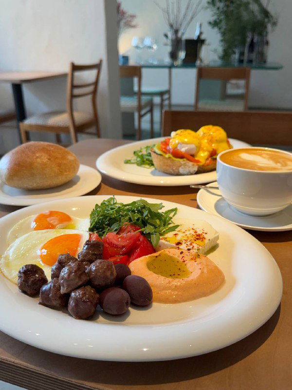 Большой греческий завтрак в Salty: тарелка с глазуньёй, маслинами, томатами, соусами, рядом булочка и чашка кофе на столе.