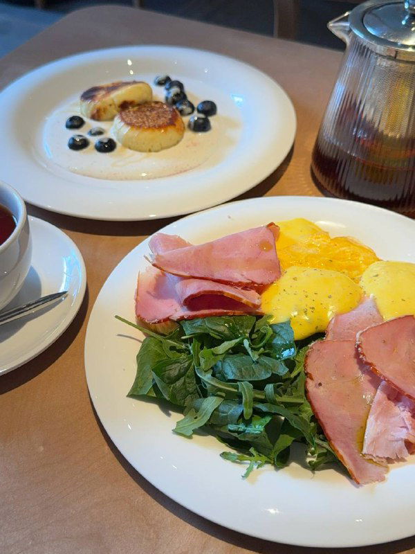 Запечённый омлет с окороком и муссом голландез на белой тарелке, рядом чайник и вторая тарелка с гарниром в кафе Salty.