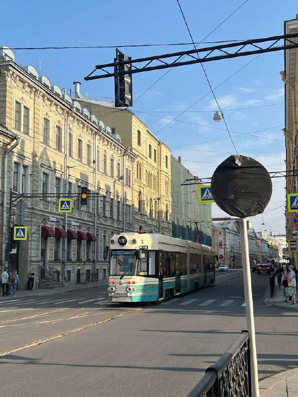 Бирюзово‑белый трамвай на широкой городской улице Петербурга под голубым небом: контактная сеть, пешеходы и городской ландшафт вдоль маршрута.