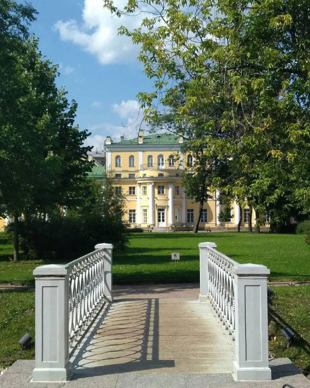 Усадьба Державина: вид на главный дом через белый деревянный мост, газон и зеленые кроны деревьев на фоне неба.