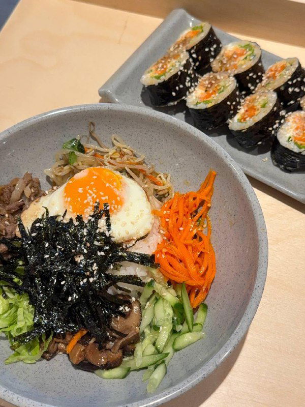 Тарелка с пибимпабом и рядом роллы кимпаб: жареное яйцо, овощи, морепродукты и рис — крупный план порции.