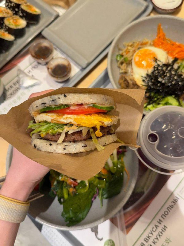 Крупный план бургера Ким: рисовые «булочки», мраморная говядина, свежие овощи и соус в бумажной упаковке для выноса.