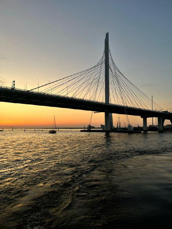 Вечерний вид на мост через залив в Петербурге: опоры моста и яхты на горизонте на фоне золотистого неба и отражения в воде.