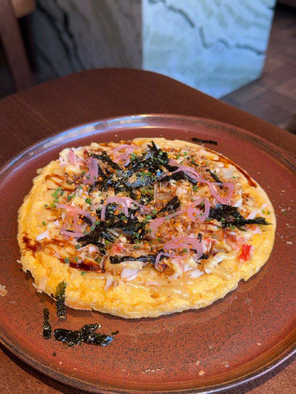 Омлет «окономияки» с крабом: круглая пышная яичная основа с крабовой начинкой, соусами и морскими водорослями на коричневой тарелке.