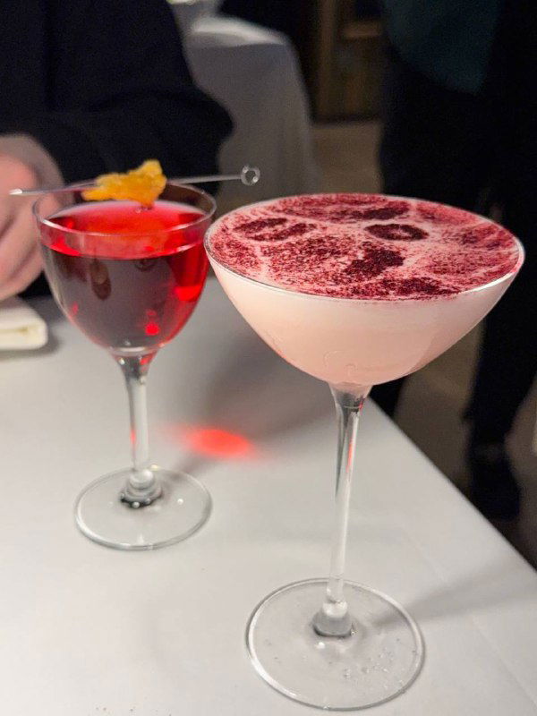 Два коктейля на столе в ресторане: розовый коктейль и насыщенно-красный напиток, аккуратная барная подача и хлебная корзина на заднем плане.
