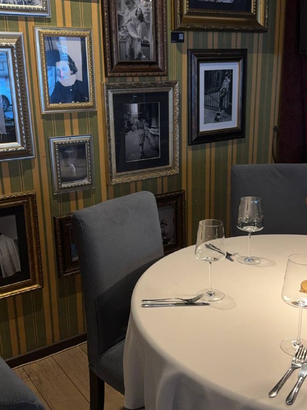 Небольшой столик у стены, украшенной множеством ретро-фотографий и рамок, сервировка для двух гостей и мягкий приглушённый свет.