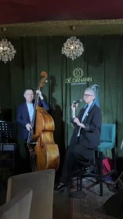 Джазовый дуэт на сцене: контрабасист и вокалист в формальной одежде на фоне зелёной занавески и логотипа заведения, живое выступление вечером.