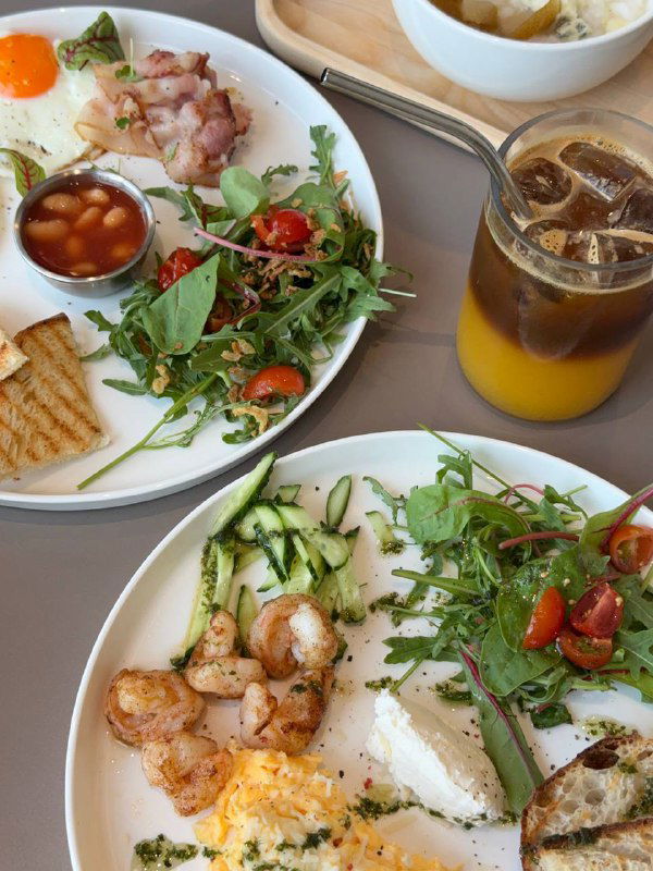 Тарелки с завтраками: зелёный салат, креветки, тосты и стакан холодного кофе — утреннее меню Kos.Place вблизи.