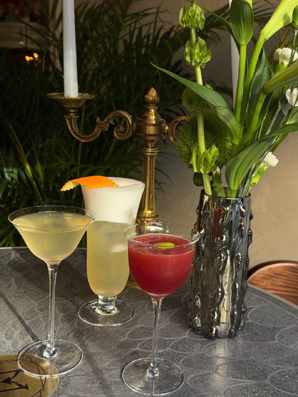 Несколько коктейлей разного цвета и формы бокалов на столе рядом с вазой и подсвечником — снимок показывает ассортимент напитков и подачу.