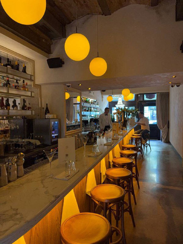 Длинная барная стойка с высокими стульями и подвесными жёлтыми светильниками, вид на рабочую зону бармена и современно-ретро оформление заведения.