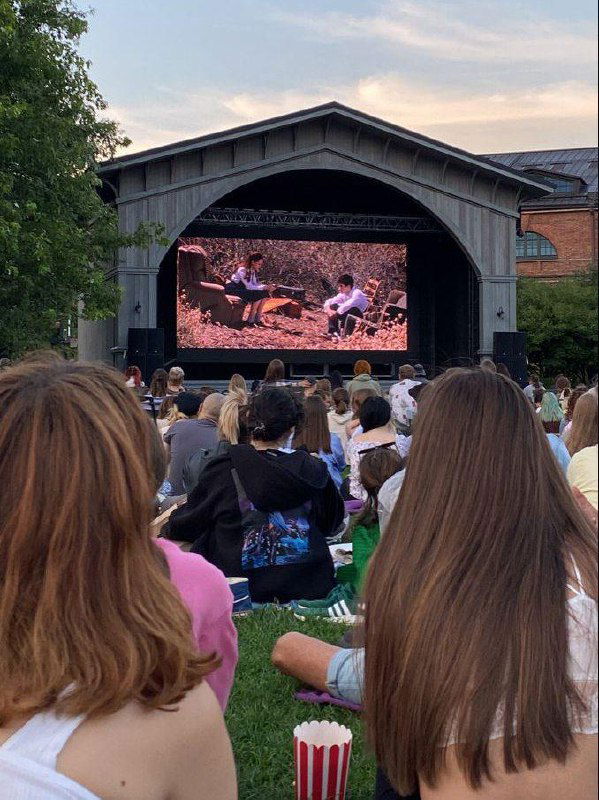 Зрители на лужайке перед летним театром: люди сидят на пледах и стульях, большой экран показывает фильм, вечернее небо над парком.
