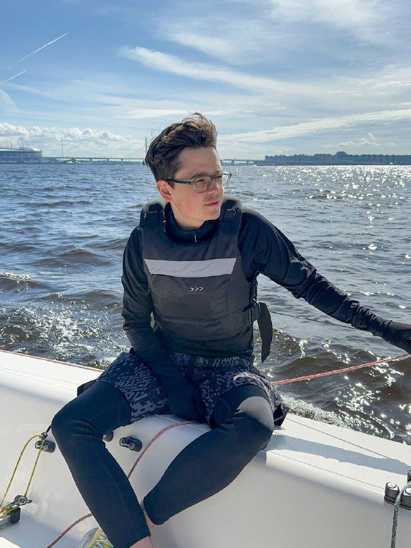 Участник тренировки в спасательном жилете сидит на борту парусника, концентрируется на управлении и наблюдает за ветром.