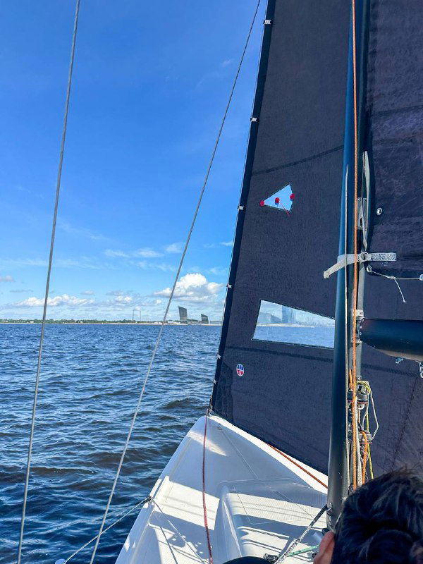 Вид с палубы на поднятый парус и мачту под голубым небом: парус готов к работе, виден горизонт залива.