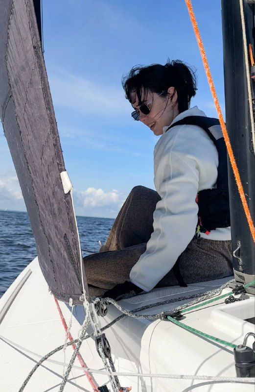 Пассажирка в жилете сидит на борту парусника, держит снасти; спокойная морская сцена во время занятия на воде.