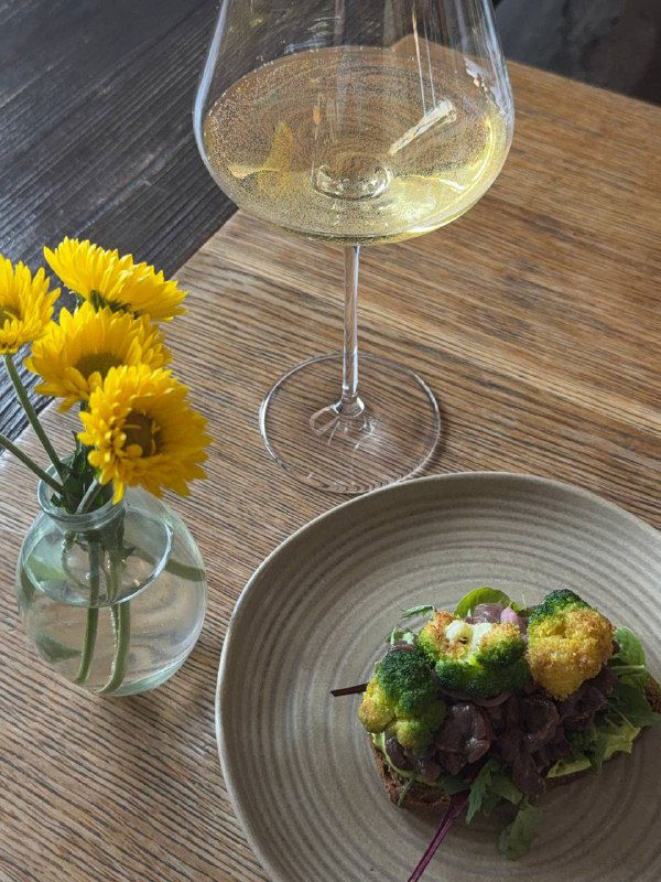 Бокал белого вина на столе рядом с вазой с желтыми цветами и аккуратно уложенной закуской на тарелке.