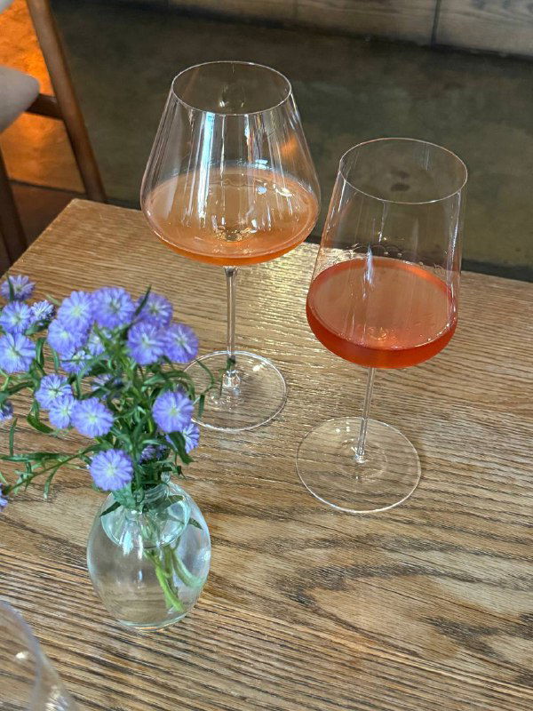 Два бокала с оранжевым и розовым винами на столе: цвет напитков, свет и маленький букет васильков в вазе.